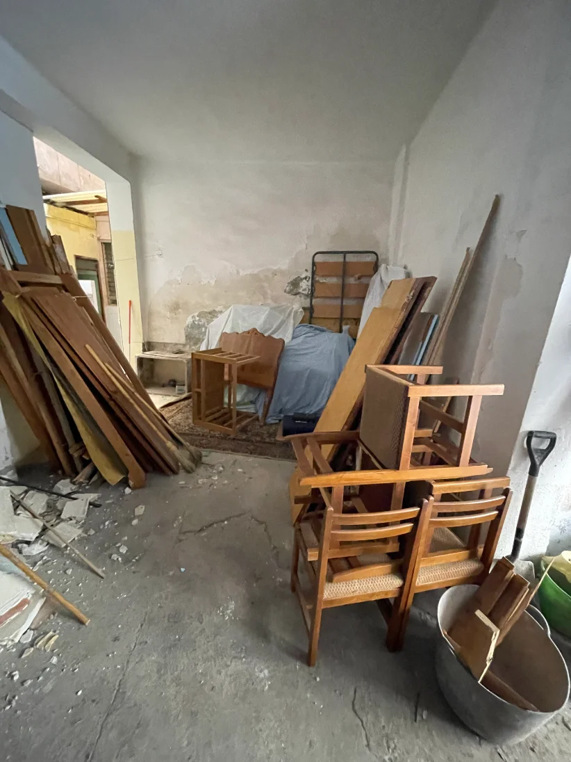 Bajo interior antes de la reforma: paredes descascarilladas, suelo de cemento y muebles viejos y tablones amontonados.