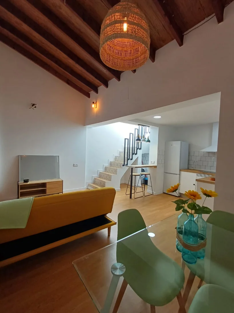 El mismo interior terminado: salón y cocina en una sola estancia, vigas de madera restauradas y escalera con peldaños de baldosa hidráulica.